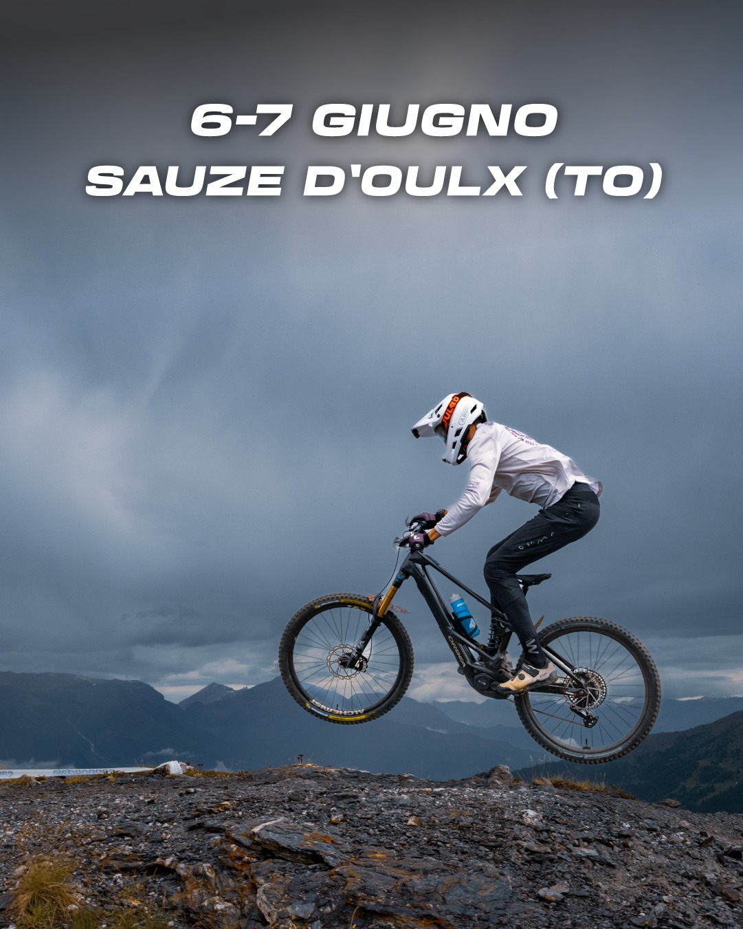 Ciclista di enduro che salta su una superficie rocciosa e sullo sfondo le montagne ed il cielo grigio 